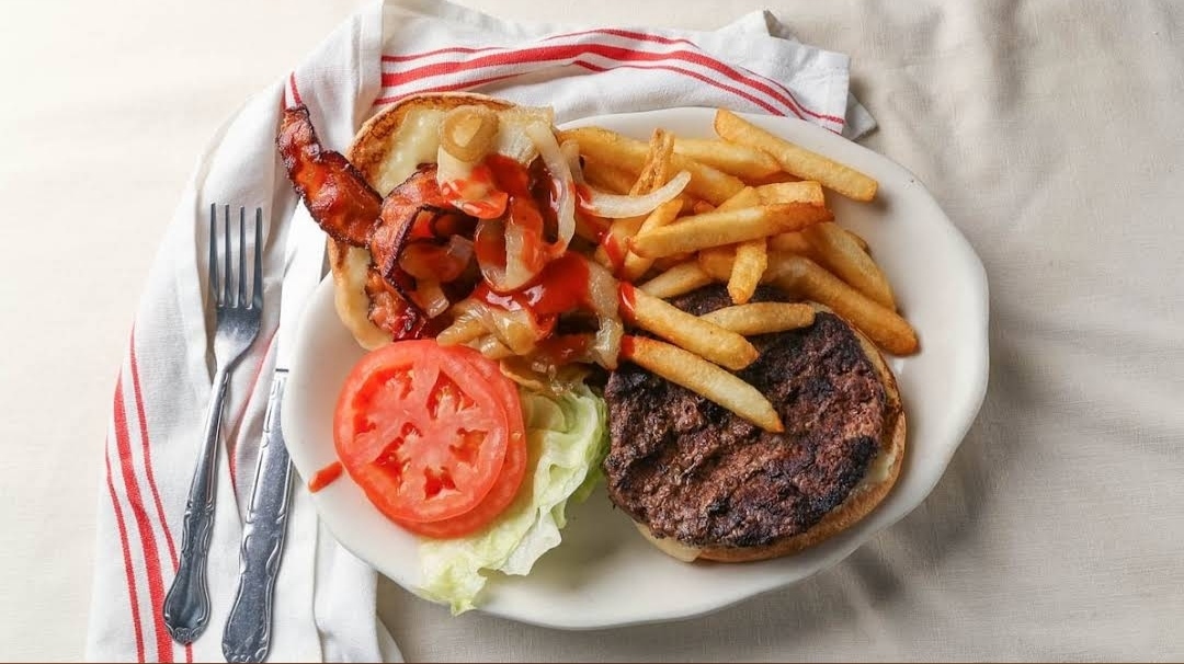 Plate with a delicious cheeseburger topped with bacon, caramelized onions, and ketchup, served with crispy french fries, lettuce, and a slice of tomato at Crosstown Diner in the Bronx