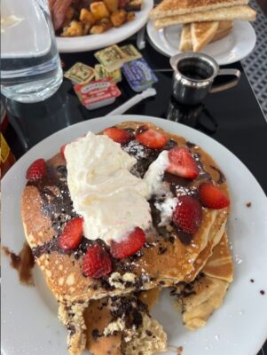 A plate of fluffy pancakes topped with whipped cream, fresh strawberries, and chocolate drizzle, accompanied by toast, breakfast potatoes, and syrup at Diner 24 in Midtown Manhattan